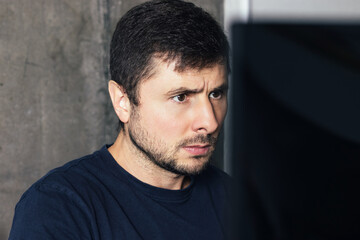 Handsome serious bearded man sitting at the office desk, working and looking at computer screen against concrete wall in modern office