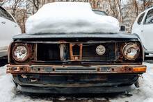 Old Abandoned Car In The Snow. Broken And Wrecked Vehicle Forgotten On Parking. Discarded Rusty Car Occupies A Parking Space