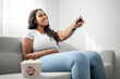 © Louis-Paul Photo - Attractive young African woman sitting on the couch at home eating popcorn