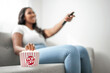 © Louis-Paul Photo - Attractive young African woman sitting on the couch at home eating popcorn