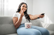 © Louis-Paul Photo - Attractive young African woman sitting on the couch at home eating popcorn