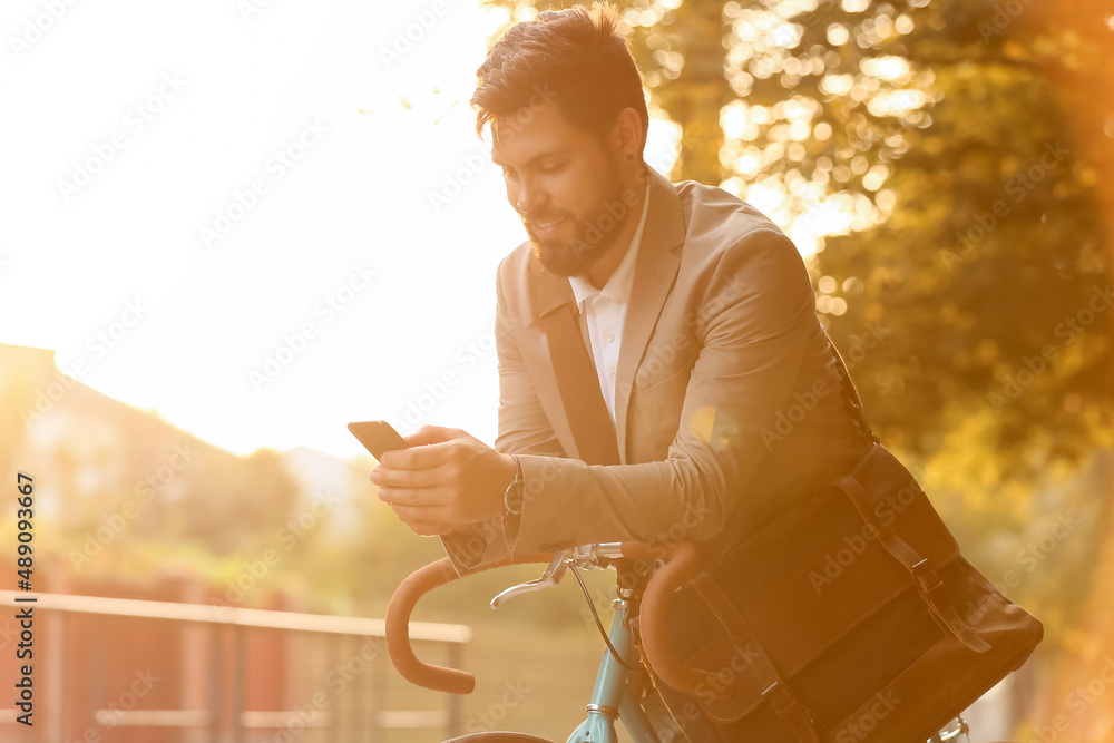 Young bearded businessman with bicycle using mobile phone on city street