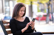 © bilanol - Young woman sitting on street bench browsing her cellphone outdoors on warm summer day. Communication and mobile connection concept