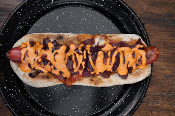 Closeup view of a delicious hot dog with crispy bacon, onions and cheddar cheese, in a black dish on the restaurant wooden table. 