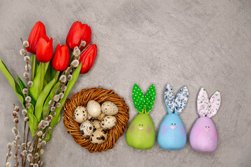 Easter decoration on the grey concrete background. Red tulips, quail eggs in the brown twig nest and colourful Easter toys shaped as a bunny. Easter decoration with the space for text.