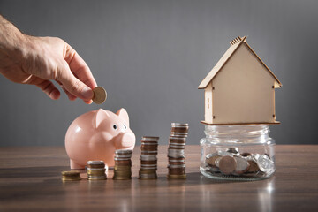 Male hand putting coin into piggy bank. House model and stack of coins on the table