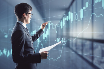 Canvas Print - Attractive young european businessman with glowing candlestick forex chart grid hologram standing in blurry office interior. Innovation, trade and finance concept. Double exposure.