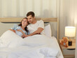 © Bangkok Click Studio - Happy Caucasian adult couple laugh and smile while talking fun and embrace each together on bed as enjoy resting under warm quilt to sleep in bedroom on romantic night at home