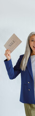 Wall Mural - Excited mature Asian model with long hoary hair in blue jacket holds gift card and Sale sign posing on light grey background in studio