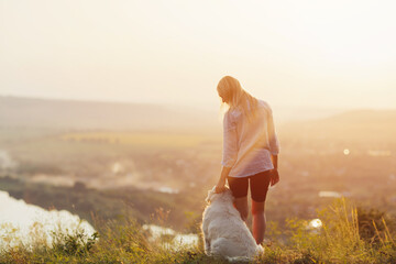 Wall Mural - Blonde woman and her dog standing on hill and enjoying amazing view over river and mountains at sunset
