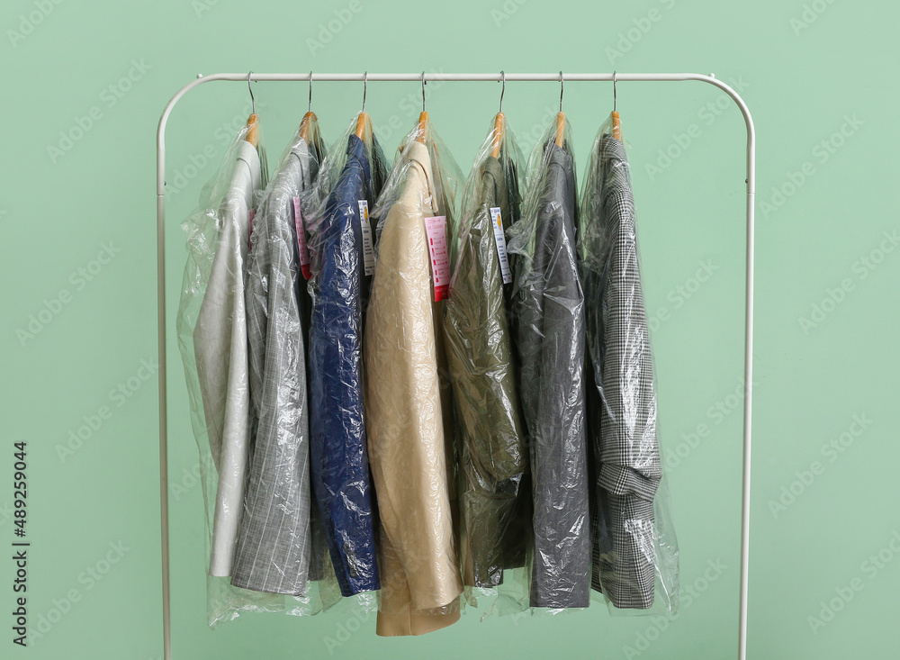 Rack with clean jackets in plastic bags on green background