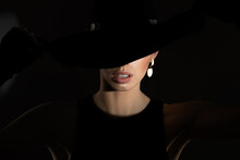 Elegant Woman In A Black Dress And Black Hat On A Dark Background.