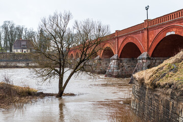 Poster - Flood in spring day. Flooded road and tree next to old red brick bridge. Kuldiga, Latvia.