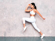 © halayalex - Fitness woman in sexy sports clothing. Young beautiful model with perfect body. Female posing in studio near grey wall at summer sunny day. Jumping and running