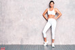 © halayalex - Fitness confident woman in white sports clothing. Sexy young beautiful model with perfect body. Female posing near grey wall in studio. Stretching out before training
