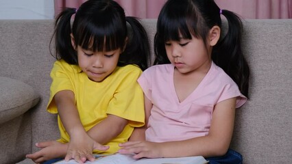 cute asian sisters reading a book lying on the sofa at home.