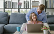 © Bangkok Click Studio - Millennial young Caucasian lover couple beautiful wife sitting on floor browsing surfing internet shopping online via laptop computer while handsome husband on cozy sofa help massaging shoulders