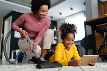 Wall Mural - Happy black mother doing household while teenager girl using digital gadgets at home.