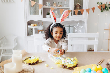 Wall Mural - Little African American girl paints Easter eggs at home in the kitchen. happy easter.