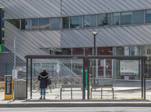 Lone Woman In Winter Coat And Baseball Cap Waiting Inside A Glass Bus Shelter, Office Building In Background