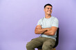 © luismolinero - Young man sitting on a chair over isolated background with arms crossed and happy