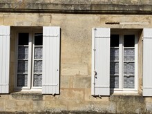 Saint Emilion, Bordeaux, France. Stone Shabby Wall With Two Windows. White Wooden Opened Shutters. Vintage White Lace Curtains