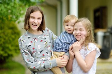 Wall Mural - Two big sisters and their toddler brother having fun outdoors. Two young girls holding baby boy on summer day. Children with large age gap. Big age difference between siblings.