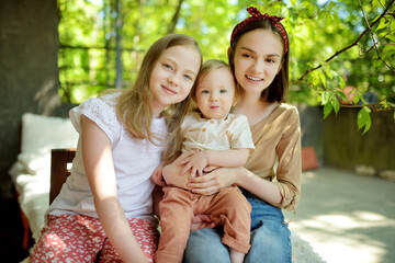 Wall Mural - Two big sisters and their toddler brother having fun outdoors. Two young girls holding baby boy on summer day. Children with large age gap. Big age difference between siblings.