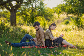 A couple is sitting on a lawn in a summer park and looking at phones