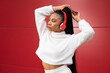 © Sergio Mena - Cheerful African American woman with long hair and headphones dressed in sportswear on a red background