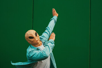 Alien doing dab posture and gesture against a green wall background. Extraterrestrial with human clothes. Concept of victory and satisfaction. Happiness and freedom immigration concept