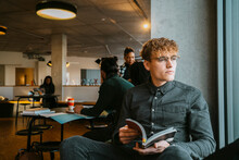 Thoughtful Young Blond Man Sitting With Book In University Cafeteria