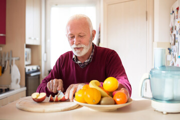 Wall Mural - Senior man eating apple and other fruits in the kitchen on a sunny morning.