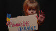 © Andrii Iemelianenko - Scared Ukrainian poor dirty toddler girl homeless protesting war conflict raises banner inscription cardboard massage Hands Off Ukraine. Crisis, no peace, stop Russian aggression. Child against war