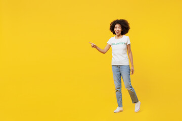 Wall Mural - Full body young woman of African American ethnicity in white volunteer t-shirt walk go point finger aside workspace isolated on plain yellow background. Voluntary free assistance help grace concept.