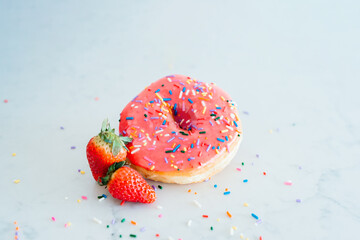 A pink frosted donut with colorful sprinkles with strawberries on a white marble background