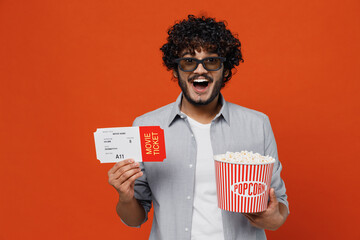 Happy jubilant exultant young bearded Indian man 20s years old wears blue shirt in 3d glasses watch movie film hold bucket of popcorn movie ticket isolated on plain orange background studio portrait.