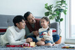 © Pruksachat - Asian young LGBTQ gay couple giving gift to little Caucasian adopted kid in living room at home. Kid smiling and delighted to receive doll as a present on birthday or special day. LGBT family concept