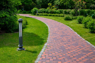 iron ground lantern garden lighting of park curved path paved with stone tiles in park among plants, evergreen bushes and foliage trees surrounded by eco friendly green lawn on sunny day, nobody.