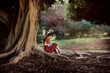 Cute european kid girl in a dress sits near a big tree in the park. A child writes poetry on large tree roots, a girl makes notes