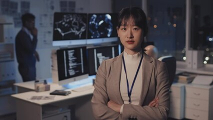 Waist-up of young Asian woman dressed formally, standing with arms crossed on chest in corporate office at night, posing and looking on camera, colleagues working on background