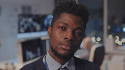 Close-up of young Black male IT specialist standing in corporate office at night, posing and looking on camera