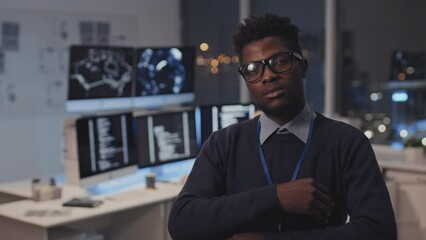 Waist-up of young Black male IT manager putting on eyeglasses, crossing arms on chest in corporate office at night, posing and looking on camera, colleagues working on background