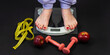 Female legs stand on an electronic scale for weight control with dumbbells and a centimeter on a dark background. The concept of sports training, diets and weight loss