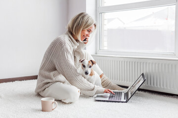 Wall Mural - Happy middle aged woman with dog using her laptop at cozy white home