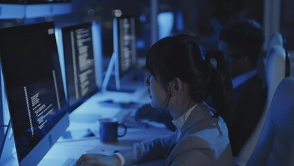 Side-view of young Asian woman sitting on chair at desk in dark modern office, using computer at night, blurred colleagues working on background