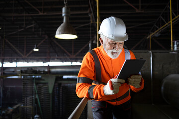 Wall Mural - Industrial engineer worker wearing safety uniform and hardhat standing on metal staircase and checking production on tablet computer. Factory interior.