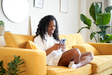 Cheerful Woman Using Mobile Phone At Home. African American Smiling Lady Is Laying On The Sofa.