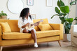 © CarlosBarquero - Young African American woman sings while playing a ukulele in the cozy living room of her home.
