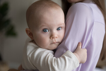 Portrait of sweet baby resting in mothers arms, looking at camera, touching mama shoulder. New mom holding little kid, embracing child with tenderness, love, care. Motherhood concept